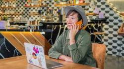 A SCORE client in an olive green jacket talks on a cell phone while sitting in front of an open laptop.